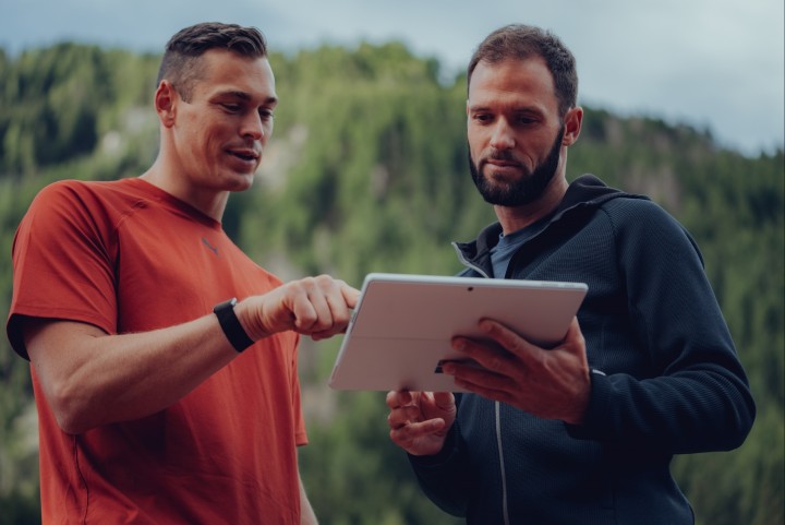 Athlet mit Uhr am Handgelenk zeigt seinem Trainer etwas auf dem Tablet, im Hintergrund Berge