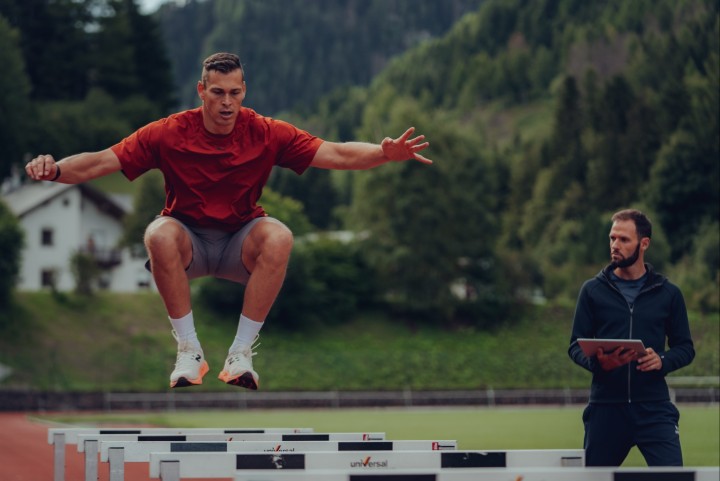 Athlete doing two-footed hurdle jumps on the track, a coach with a tablet beside him