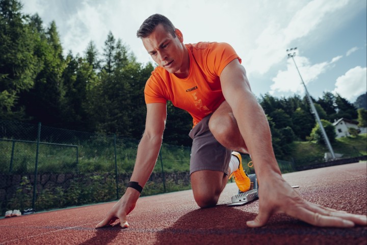 Athlete in a low starting position on the track, trees and a floodlight mast in the background