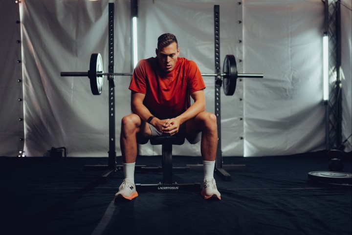 Athlete sitting on a bench in a weight room in front of a barbell, hands clasped, looking down