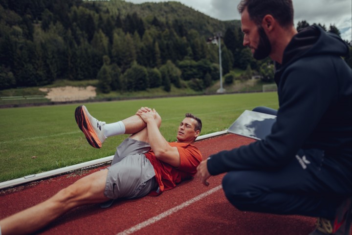 Coach kneeling next to an athlete who lies on the track and pulls one knee to his chest