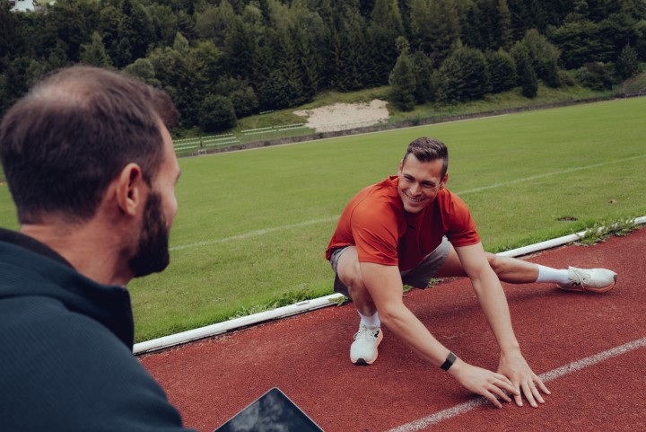 Trainer mit Tablet im Vordergrund, dahinter dehnt sich ein lächelnder Athlet auf der Laufbahn
